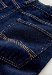 Granatowy jeansowy materiał z widocznym przeszyciem w jasnobrązowym kolorze. Posiada zaokrągloną tylną kieszeń i teksturowaną powierzchnię.