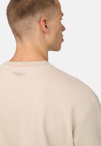 Beige garais piedurkņu krekls ar strukturētu audumu un apaļu izgriezumu. Aizmugurējā apkaklē redzams neliels etiķete ar tekstu "TECH".