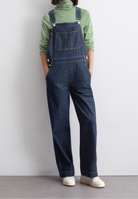Denimové overal s nastavitelnými ramínky, přední kapsou, širokými nohavicemi a tmavě modrou barvou. Nositelný přes zelený a bílý pruhovaný rolák.
