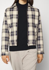 Veste à carreaux présentant des couleurs crème et marine, avec fermeture éclair et un design à col, accompagnée d'un col roulé noir et de pantalons clairs.