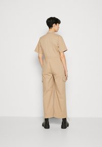 Beige utility jumpsuit with short sleeves, cargo pockets, and a relaxed fit. Made of durable cotton blend, features a drawstring at the hem.