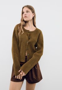 Cardigan côtelé vert olive avec des boutons, un col rond et des manches longues. Porté sur un short marron, mettant en avant un design court.