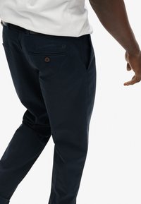 Marineblaue Baumwollhosen im Slim-Fit-Stil mit Gesäßtaschen und einem braunen Knopfdetail. Der Stoff hat eine glatte Textur.