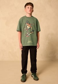 Zelena pamučna majica s grafikom žabe, crne terenske hlače s bočnim džepovima i zelene tenisice s bijelim detaljima. Jednostavan dizajn, opušteni kroj.