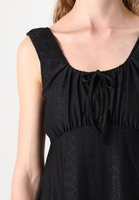 Vestido preto sem mangas com decote franzido, detalhe de laço e tecido texturizado com padrões verticais e em diamante.