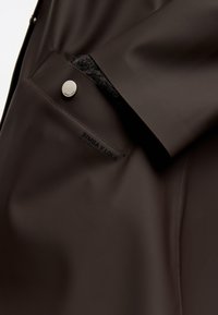 Abrigo marrón impermeable con textura suave, que presenta un botón a presión y detalle del logo en el bolsillo. La costura lateral es elegante y ajustada.