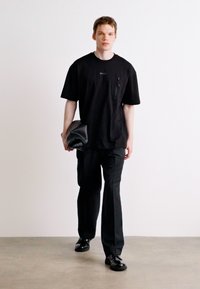 Černé oversized tričko s kapsou, spárované s černými kalhotami a černými botami. Modelka drží černou koženou kabelku.