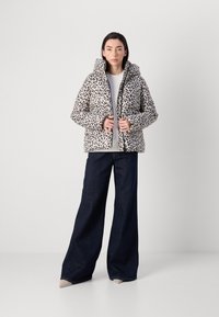 Eine Frau in einer Leopard-Print-Pufferjacke, weißem Shirt und dunklen, weiten Jeans steht vor einem hellgrauen Hintergrund und hält die Jacke mit beiden Händen.