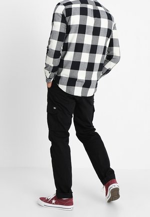 Homme portant une chemise à carreaux noir et blanc, un pantalon cargo noir et des baskets rouges marchant de dos les mains dans les poches sur fond blanc.