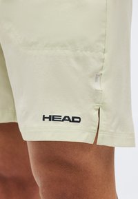 Μπεζ αθλητικό σορτς με μικρό πλαϊνό σκίσιμο, μαύρο λογότυπο "HEAD" κοντά στο κάτω μέρος, φορεμένο σε μηρό με καστανό δέρμα.