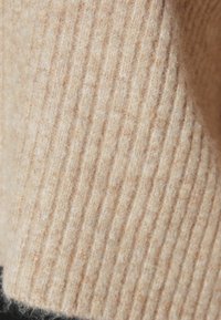 Beigea ribbineulottu kangas, jossa on pehmeä tekstuuri; siinä on pystysuorat linjat ja lämmin sävy, joka luo kodikkaan ja kutsuvan ulkonäön.