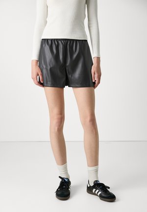 Mujer con pantalones cortos de cuero negro, top blanco de manga larga, calcetines blancos y zapatillas Adidas negras de pie contra un fondo blanco liso.