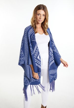 Blauwe sjaal met een patroon van witte geometrische ontwerpen, met rafelige randen, gemaakt van een zachte stof, gedragen over een witte top en gecombineerd met een witte broek.