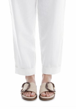 Sandalias beige tejidas con una banda ancha, con un detalle marrón y hebilla, usadas con pantalones blancos recortados, mostrando un diseño sencillo y casual.