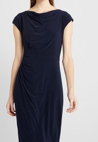 Vestido azul marino hecho de un tejido suave y drapeado, con mangas de capucha, un escote drapeado y detalles fruncidos en el lateral.