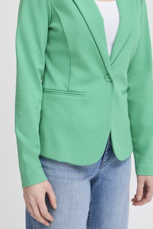 Personne portant un blazer vert par-dessus un top blanc avec un jean bleu clair, mains détendues le long du corps, sur un fond uni.