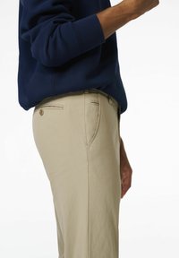 Beige katoenen broek met een slim fit, zijzakken en een achterzak met drukknopsluiting. Draagt met een marineblauwe sweatshirt.