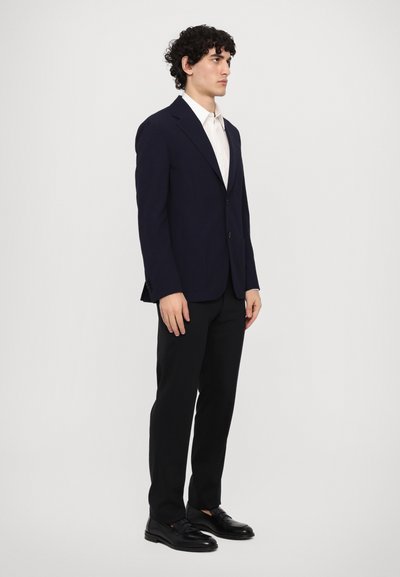 Blazer blu navy con rever a punta, bottone singolo e due tasche frontali, abbinato a una camicia bianca con colletto e pantaloni neri. Mocassini neri.