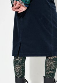 Námořnická kordrová sukně se bočním rozparkem, doplněná zelenými krajkovými legínami a černými kotníkovými botami, zdůrazňující texturu a vrstvený design.