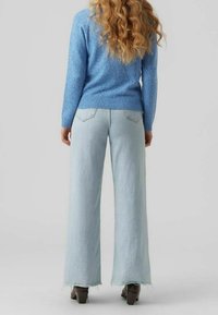 Kvinna med långt lockigt hår som bär en blå tröja, ljusblå vida jeans med fransade fållar och mörka klackstövlar, sedd bakifrån.