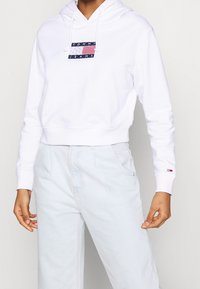 Bílá zkrácená mikina s kapucí na šňůrku. Na hrudi se nachází modro-červené logo "TOMMY JEANS". Doplněno o světlé modré kalhoty s vysokým pasem.