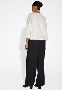 Frau mit geflochtenem Haar, die ein cremefarbenes Oberteil mit Dreiviertelärmeln, weit geschnittene schwarze Hosen und schwarze Absatzschuhe trägt, mit dem Rücken zur Kamera.