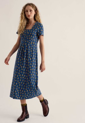 Frau mit langen, lockigen Haaren, die ein blaues, kurzärmliges, knöchellanges Blumenkleid und braune Stiefel trägt, geht vor einem schlichten Hintergrund.