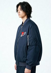 Giubbotto bomber blu navy con chiusura a zip, polsini a coste e tasca laterale. Presenta ricami di un cuore rosso e una spilla di sicurezza sul petto.