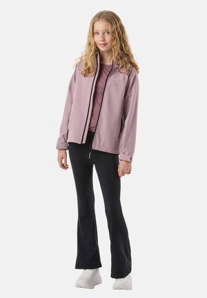 Ung person med langt blondt hår iført en mauve lynlåsjakke, mauve top, sorte udvidede bukser og hvide sneakers på en hvid baggrund.