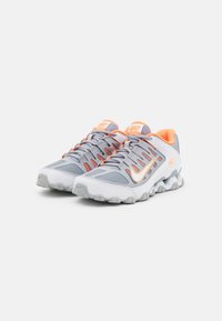 Un par de zapatillas de entrenamiento Nike en blanco y gris con acentos naranjas y suela en zigzag, colocadas una al lado de la otra sobre un fondo blanco.