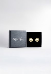 Guldaccentuerade örhängen visade i en texturerad svart smyckeslåda med innervelour, som visar två runda grönaktiga pärlor.