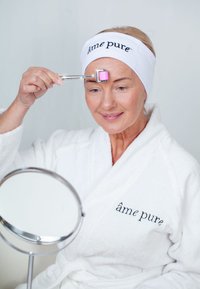 Gulltonet ansiktsruller med rosa ende holdt mot pannen av en kvinne i hvit badekåpe og hårbånd, mens hun holder et rundt speil.