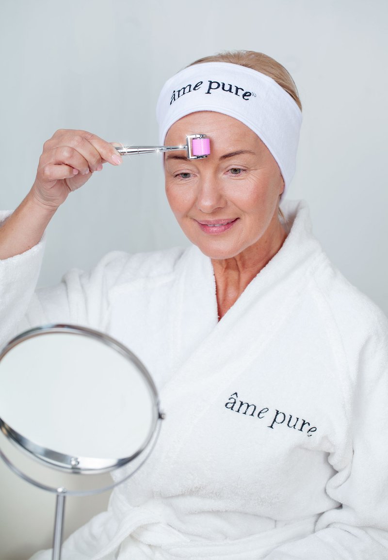 Gulltonet ansiktsruller med rosa ende holdt mot pannen av en kvinne i hvit badekåpe og hårbånd, mens hun holder et rundt speil.