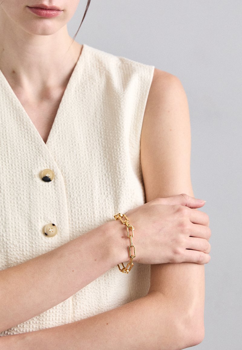 Zlatna verižica z pravokotnimi povezavami, nošena na svetli koži na roki proti teksturirani svetlo kremni bluzici z gumbi.