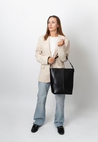 Femme portant un blazer beige, une chemise blanche, un jean clair, des mocassins noirs, tenant un grand sac cabas en cuir noir sur fond blanc.