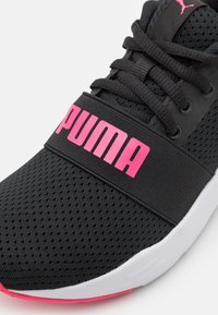 Zwarte sportieve schoen met een bovenwerk van mesh, voorzien van een opvallend roze "PUMA"-logo op een band, witte zool met roze accenten en gestructureerde oppervlakken.