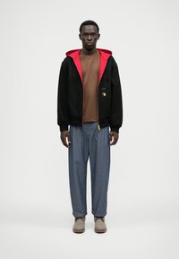 Črna kapuca jakna z rdečo podlogo, rjav majica, široke modre kavbojke in svetlo sivi škornji z gumijastim podplatom.