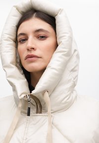 Vadderad krämfärgad jacka med en stor, strukturerad huva, med framdragkedja och beige dragsnören. Slätt, glänsande tyg med kviltat mönster.