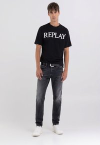 Camiseta de algodón negra con el logo "REPLAY" en blanco, combinada con jeans negros desgastados y zapatillas blancas. El modelo está de pie frente a un fondo claro.