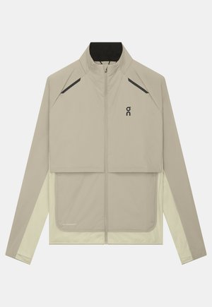 Veste beige clair zippée intégralement avec col montant, logo noir sur la poitrine, détails diagonaux noirs près des épaules et manches beige clair contrastantes.