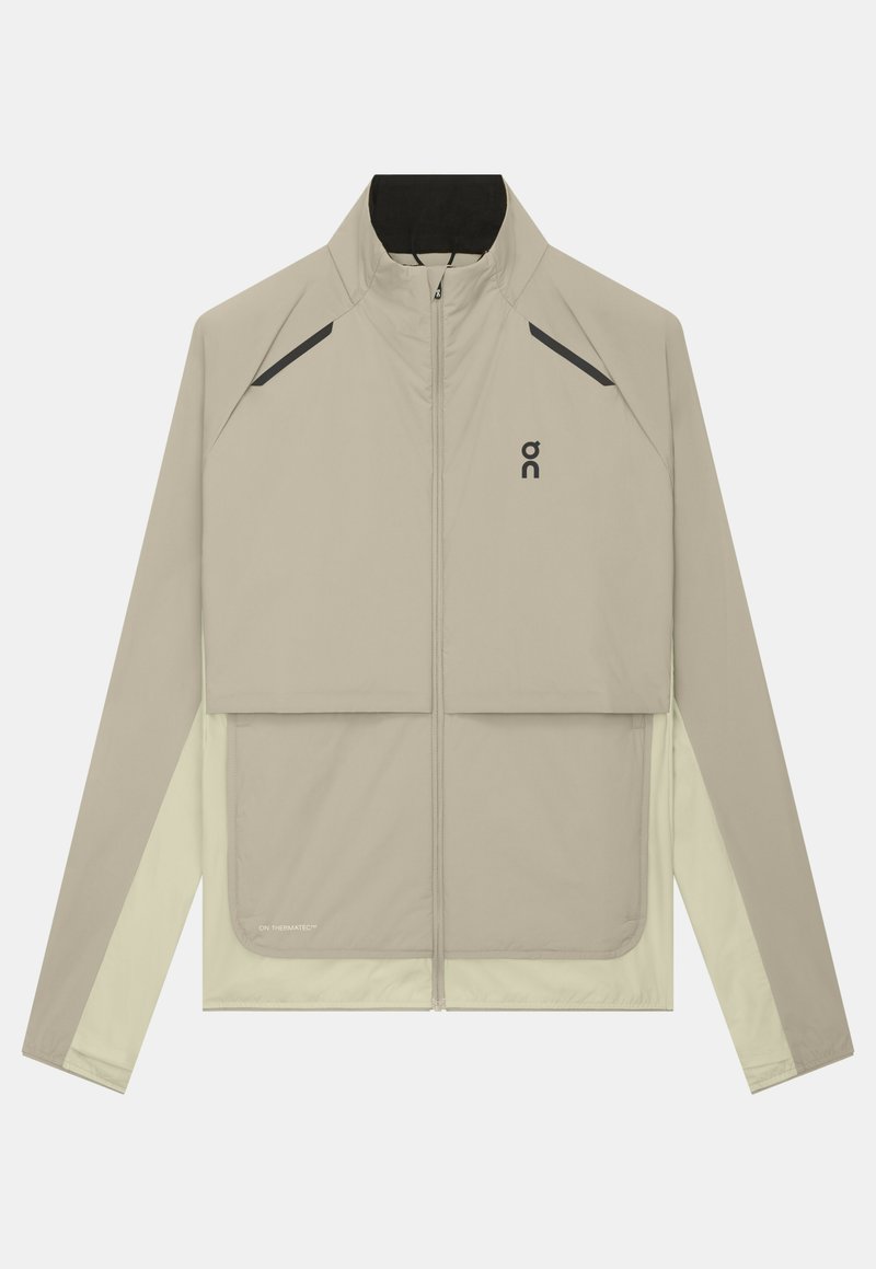 Veste beige clair zippée intégralement avec col montant, logo noir sur la poitrine, détails diagonaux noirs près des épaules et manches beige clair contrastantes.