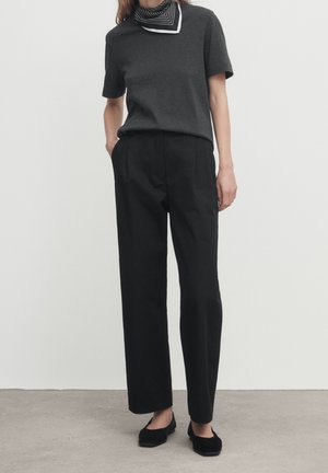 Frau trägt ein dunkelgraues kurzärmeliges Oberteil, schwarze hoch taillierte gerade Hosen, schwarze Ballerinas und ein schwarz-weiß gestreiftes Halstuch.