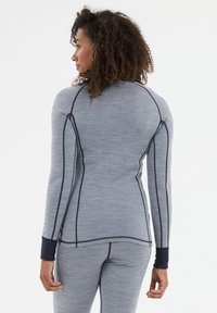 Nainen, jolla on kiharat hiukset, pukeutuneena harmaaseen, tiukkaan pitkähiharaiseen paitaan ja housuihin, joissa on mustia sauman yksityiskohtia. Hän on kääntynyt hieman sivulle valkoista taustaa vasten.