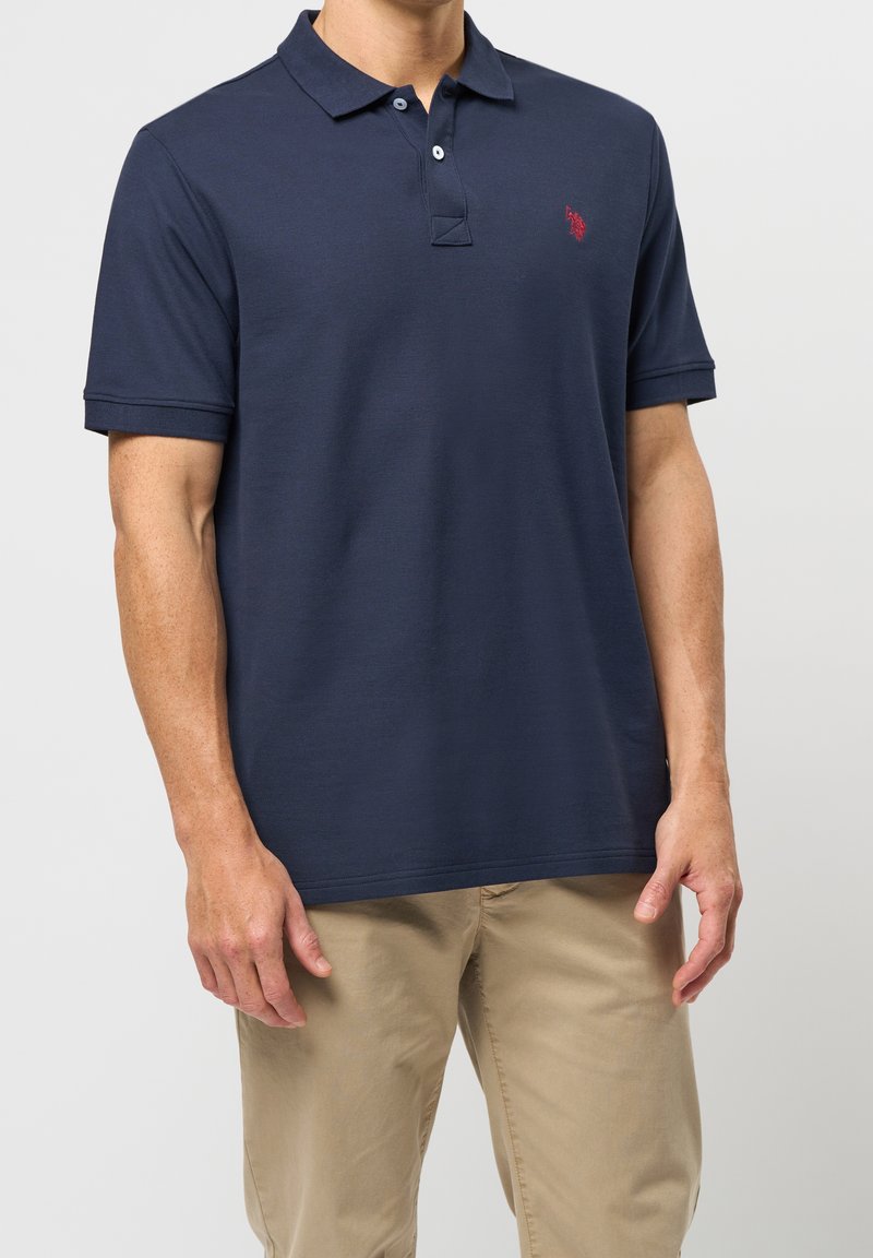 Homme portant un polo bleu marine à manches courtes et un pantalon beige, debout devant un fond uni.