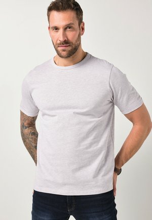 Mężczyzna z brodą i wytatuowanym ramieniem, ubrany w jasnoszarą koszulkę z krótkim rękawem i ciemne dżinsy, stoi z rękami za plecami.