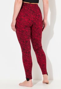 Leggings rossi con un motivo floreale nero, caratterizzati da un design aderente e un tessuto testurizzato. Accenti neri ai polsini delle caviglie.