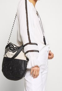 Czarnobiała torebka z misternym haftem kwiatowym, teksturowana skóra, regulowany pasek i zamek błyskawiczny; noszona na ukos.