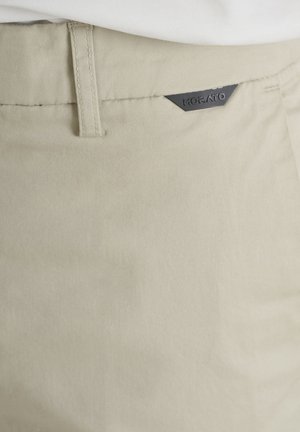Zone de la taille d'un pantalon beige montrant une passants de ceinture et une petite étiquette triangulaire noire avec le nom de la marque Korato.