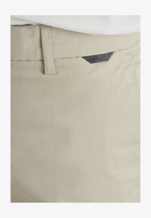 Zone de la taille d'un pantalon beige montrant une passants de ceinture et une petite étiquette triangulaire noire avec le nom de la marque Korato.