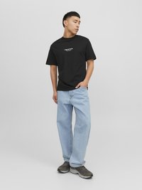 Schwarzes Baumwoll-T-Shirt, lockere Passform mit weißem Logo auf der Vorderseite. Hellblaue Jeans im geraden Schnitt, leicht verwaschen. Dunkle Sneaker.